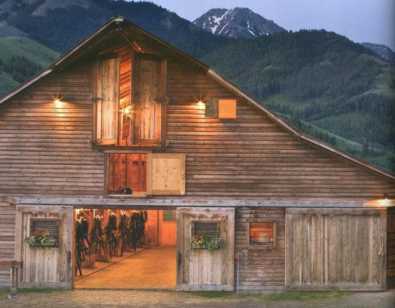 A rustic wooden barn located in a mountainous area, featuring several doors, a large peaked roof, and decorative flower boxes at the entrance.